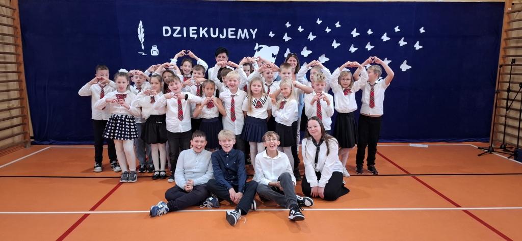 Uczniowie i opiekunowie w grupie po występie uśmiechają się i unoszą ręce w geście wdzięczności, tworząc z dłoni serduszka