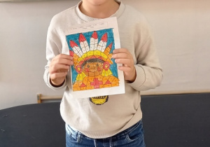 Uczeń z klasy "0" pokazuje swoją pracę z tematu "Thanksgiving", praca została wykonana według instrukcji - pomalowania danego miajsca określonym kolorem. Praca pokazuje indianina - postać, związaną z historią Święta Dziękczynienia.