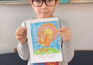 Uczeń klasy "0" pokazuje swoja pracę z tematu "Thanksgiving", praca została wykonana według instrukcji - pomalowania danego miejsca określonym kolorem. Praca pokazuje indyka - symbol Święta Dziękczy