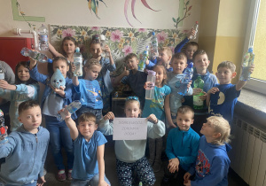 zdjęcie przedstawia uczniów klasy pierwszej, ubranych na niebiesko, trzymających w rękach butelki z wodą oraz plakat z napisem Dobra woda zdrowia doda