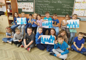 zdjęcie przedstawia uczniów klasy pierwszej, ubranych na niebiesko, trzymających w rękach narysowane kropelki wody oraz plakaty dotyczące oszczędzania wody
